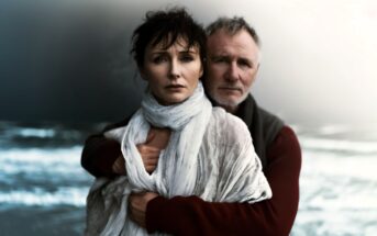 A woman wrapped in a white scarf stands in front of a man who embraces her from behind. Both look serious and pensive. The background is a blurred, moody seascape with waves and cloudy skies.
