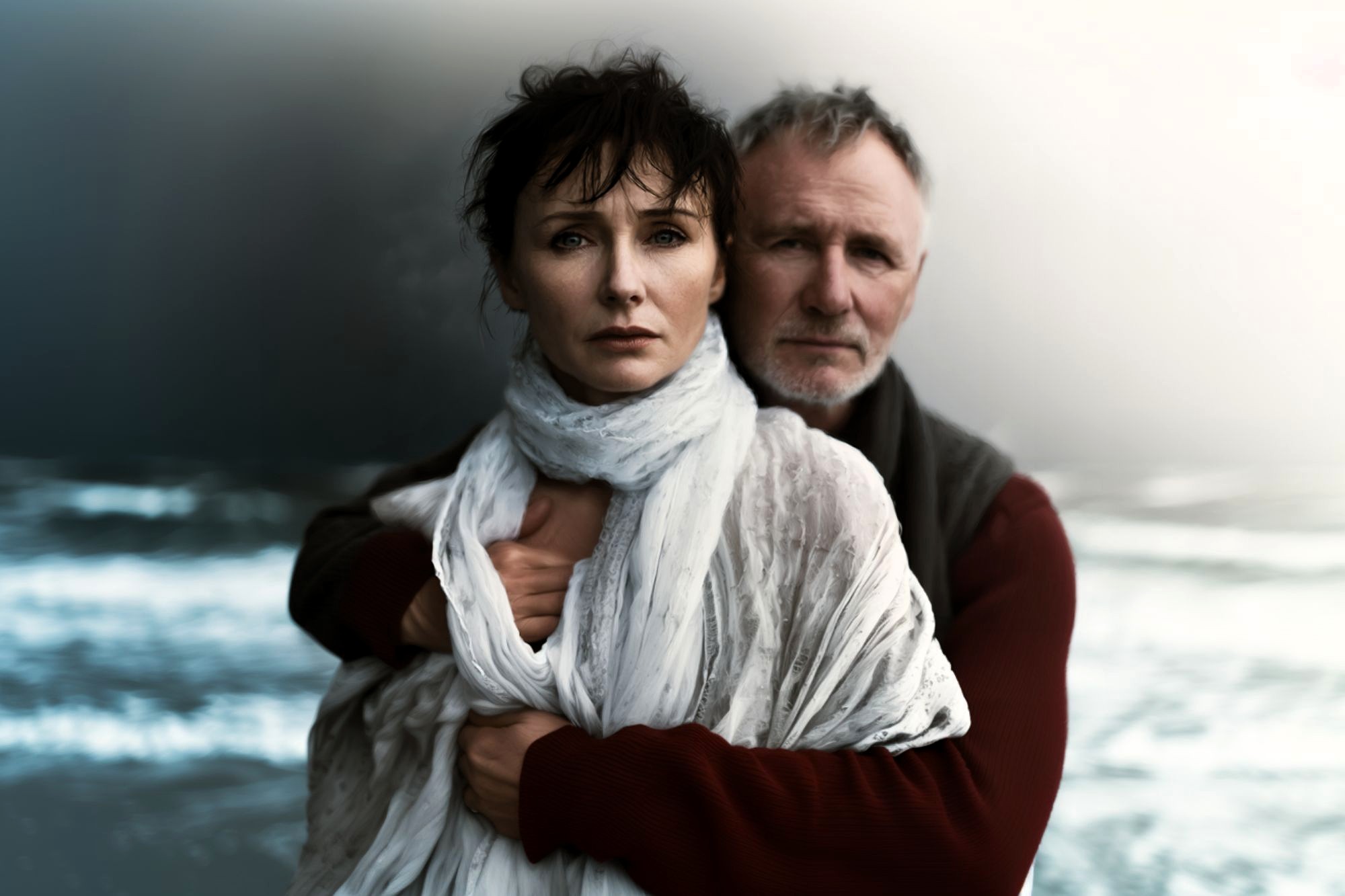 A woman wrapped in a white scarf stands in front of a man who embraces her from behind. Both look serious and pensive. The background is a blurred, moody seascape with waves and cloudy skies.