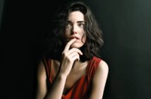 A woman with wavy dark hair and wearing a red sleeveless top gazes upward thoughtfully, with her finger resting on her lips, set against a dark background.