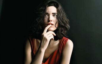 A woman with wavy dark hair and wearing a red sleeveless top gazes upward thoughtfully, with her finger resting on her lips, set against a dark background.