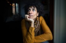 A woman with curly hair, wearing a mustard yellow top, patterned scarf, and hoop earrings, rests her chin on her hand while gazing thoughtfully out of a window in a dimly lit room.