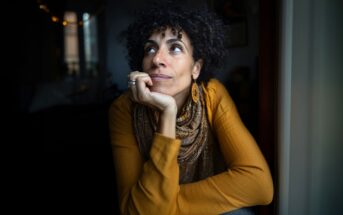 A woman with curly hair, wearing a mustard yellow top, patterned scarf, and hoop earrings, rests her chin on her hand while gazing thoughtfully out of a window in a dimly lit room.