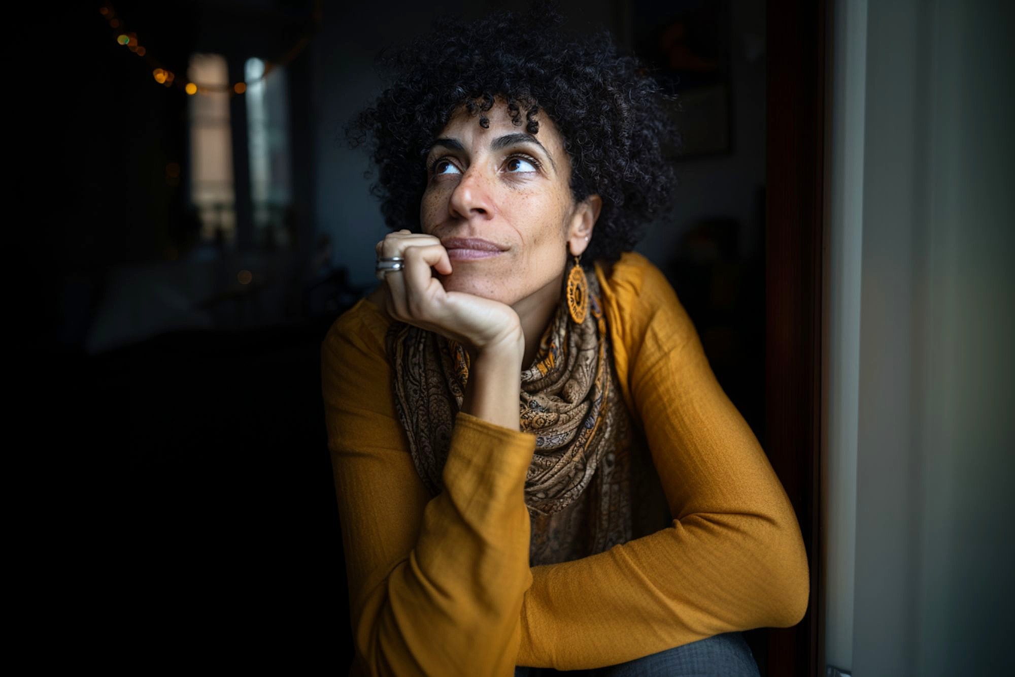 A woman with curly hair, wearing a mustard yellow top, patterned scarf, and hoop earrings, rests her chin on her hand while gazing thoughtfully out of a window in a dimly lit room.