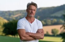 A middle-aged man with graying hair and stubble standing with his arms crossed outdoors.