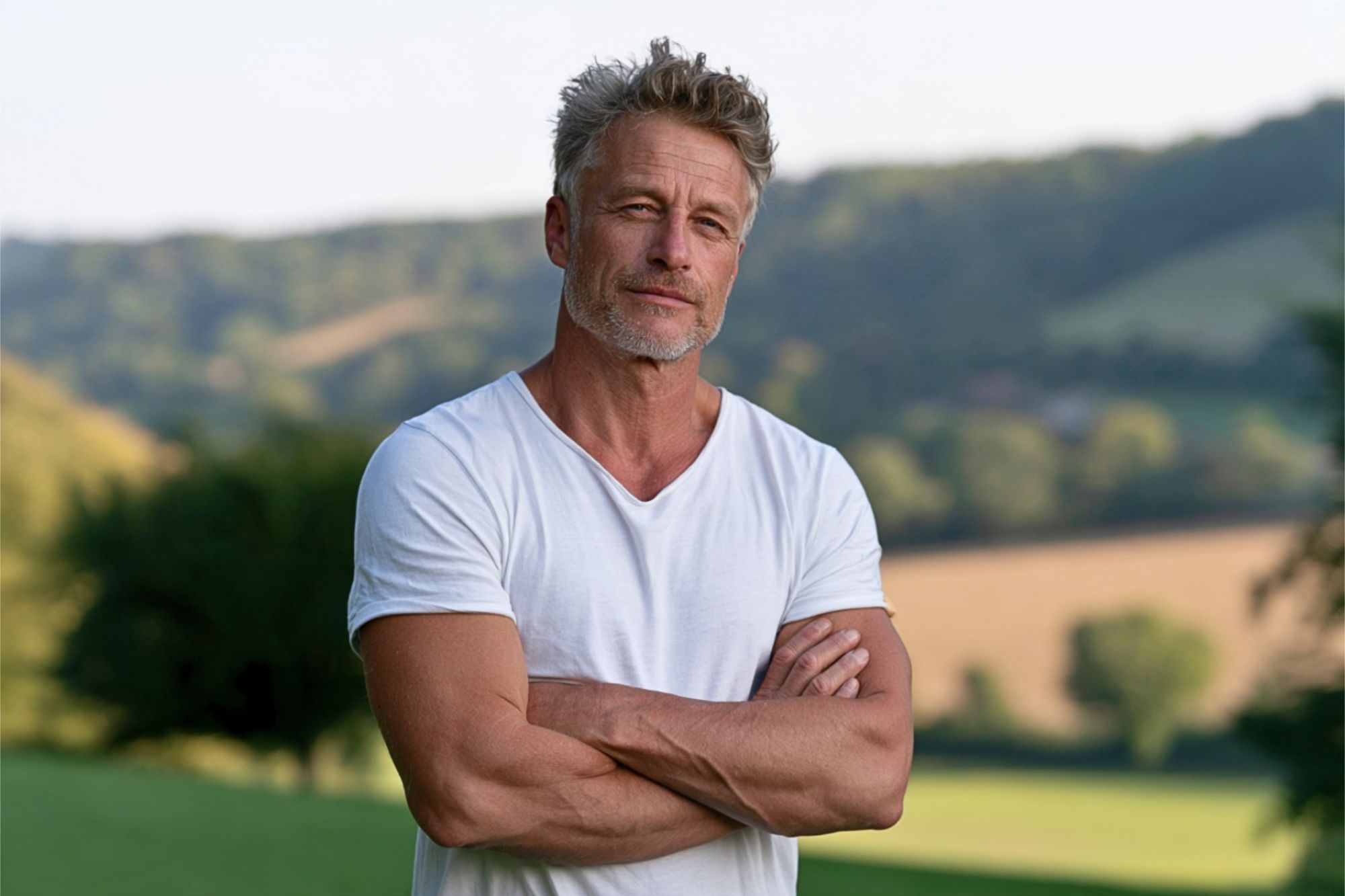 A middle-aged man with graying hair and stubble standing with his arms crossed outdoors.