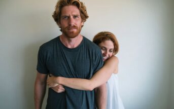 A woman with ginger hair hugs a ginger-bearded man from behind.