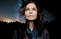 Portrait of a woman looking upward against a background of a starry night sky and silhouettes of trees.