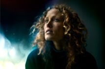 A woman with curly hair gazes upward, illuminated by soft, ethereal light against a dark, blurred background, creating a dreamy and contemplative atmosphere.