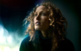 A woman with curly hair gazes upward, illuminated by soft, ethereal light against a dark, blurred background, creating a dreamy and contemplative atmosphere.