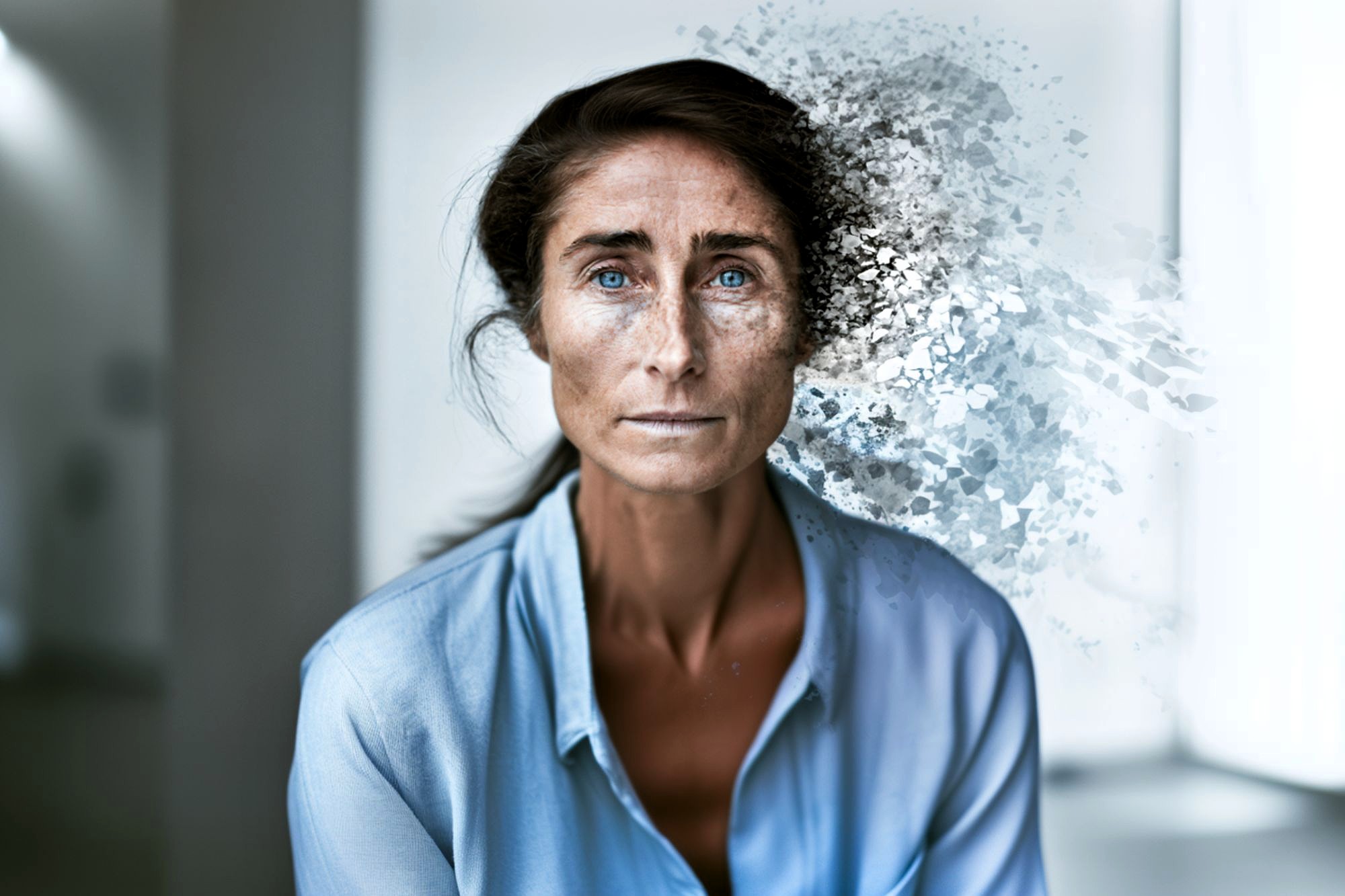 A woman in a blue shirt stares ahead as the right side of her face and hair dissolve into abstract particles, blending with a blurred indoor background.