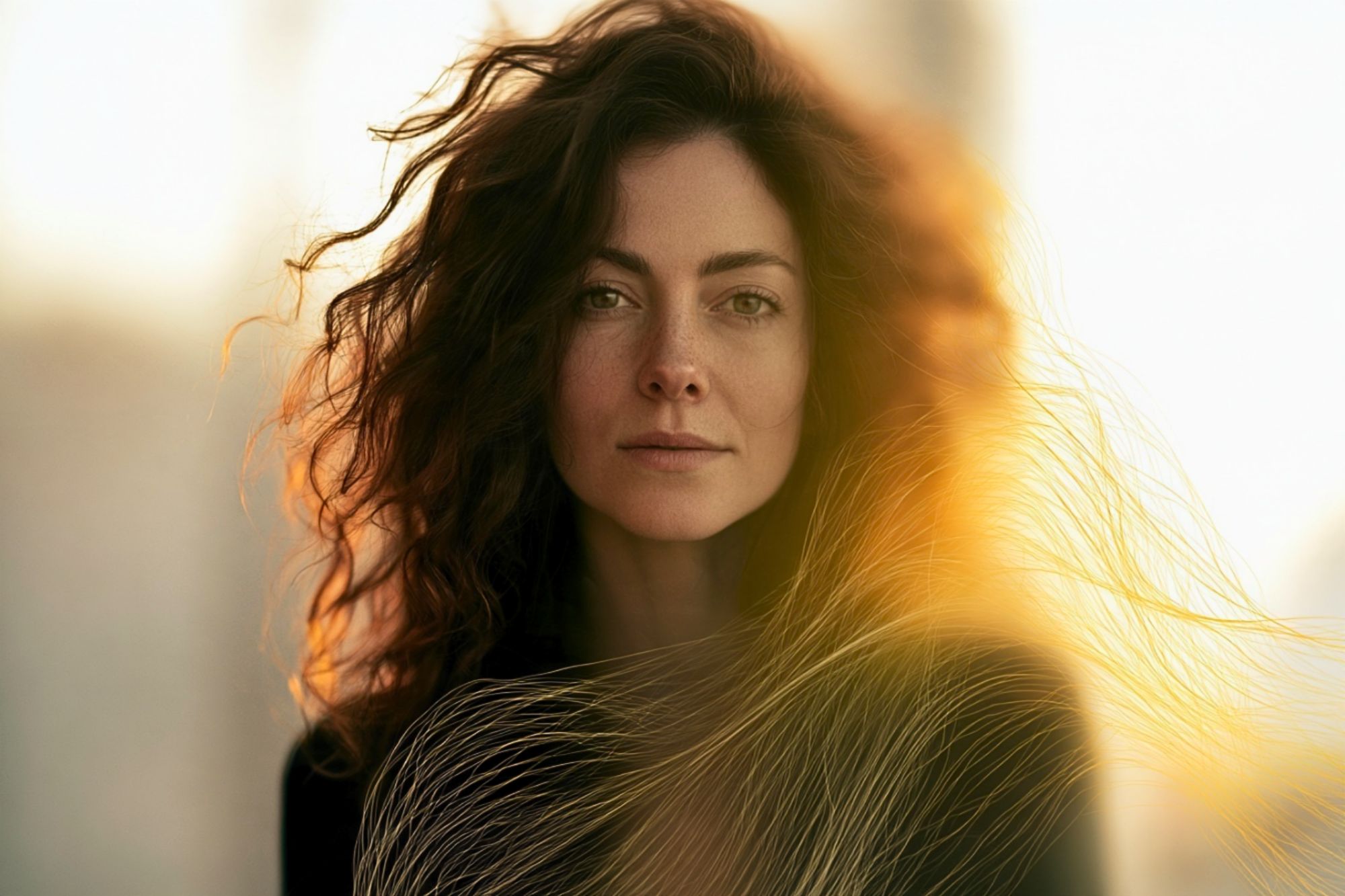 Close-up portrait of a woman with long, dark, wavy hair backlit by golden sunlight.