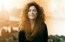 A woman with curly hair and a scarf smiles subtly, standing outdoors in soft, golden sunlight with an out-of-focus cityscape behind her.