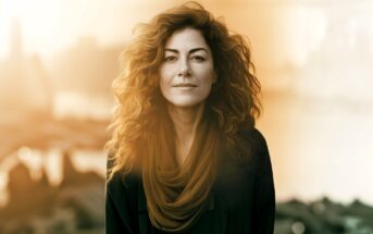 A woman with curly hair and a scarf smiles subtly, standing outdoors in soft, golden sunlight with an out-of-focus cityscape behind her.