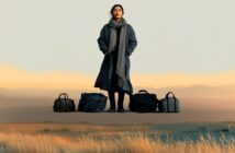 A woman in a long coat and scarf stands outdoors on dry grass with five black travel bags lined up in front of her, against a backdrop of soft hills and a pale sky.