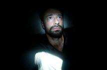A man with a beard sits in darkness, illuminated by a beam of light. He looks slightly upward with a worried or thoughtful expression, while most of his surroundings remain in deep shadow.