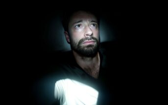 A man with a beard sits in darkness, illuminated by a beam of light. He looks slightly upward with a worried or thoughtful expression, while most of his surroundings remain in deep shadow.