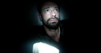 A man with a beard sits in darkness, illuminated by a beam of light. He looks slightly upward with a worried or thoughtful expression, while most of his surroundings remain in deep shadow.