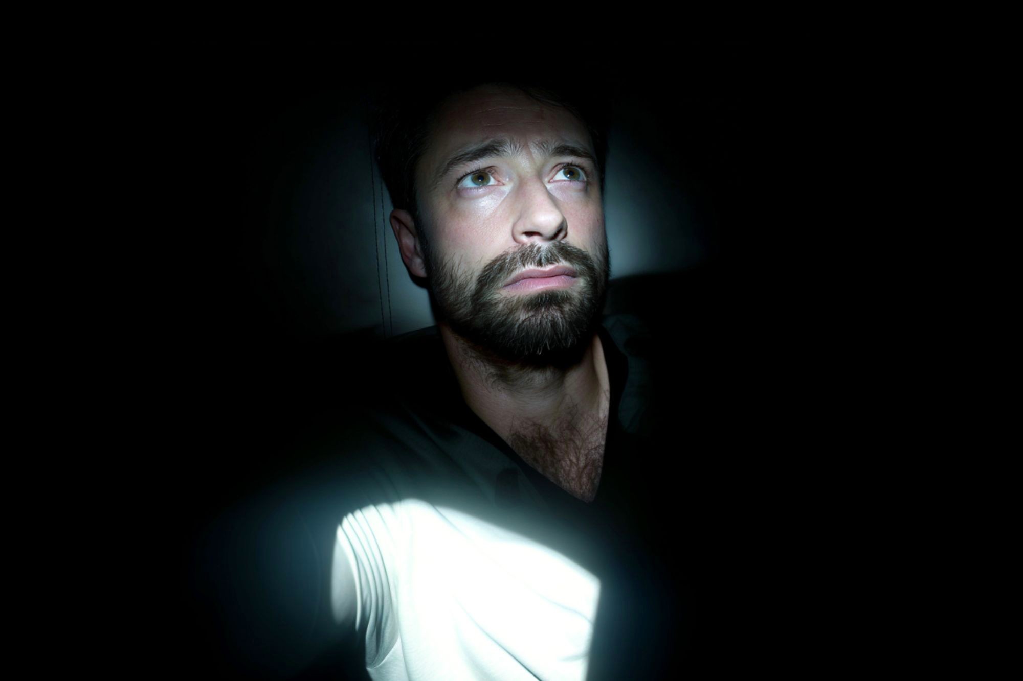 A man with a beard sits in darkness, illuminated by a beam of light. He looks slightly upward with a worried or thoughtful expression, while most of his surroundings remain in deep shadow.