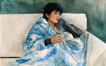 A person with short dark hair sitting on a couch, wrapped in a blue patterned blanket and holding a steaming mug.