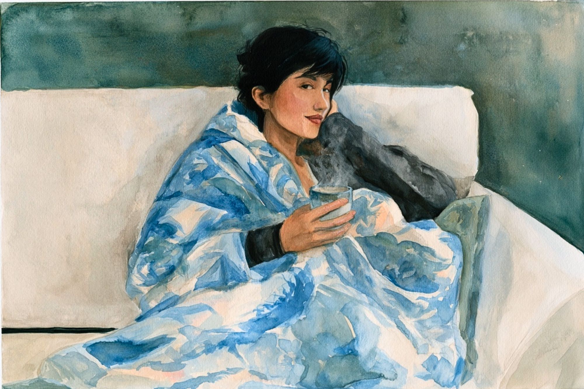 A person with short dark hair sitting on a couch, wrapped in a blue patterned blanket and holding a steaming mug.