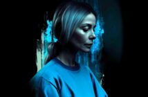 A woman with light hair in a ponytail, wearing a blue sweatshirt, faces left in a moody, blue-lit setting with abstract, dark and teal paint strokes in the background.