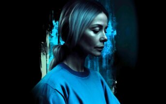 A woman with light hair in a ponytail, wearing a blue sweatshirt, faces left in a moody, blue-lit setting with abstract, dark and teal paint strokes in the background.