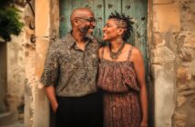 A smiling couple stands close together in front of an old, weathered blue door, looking at each other warmly. The man wears glasses and a patterned shirt, while the woman wears a sleeveless dress and large necklace.
