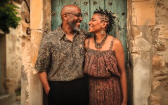 A smiling couple stands close together in front of an old, weathered blue door, looking at each other warmly. The man wears glasses and a patterned shirt, while the woman wears a sleeveless dress and large necklace.