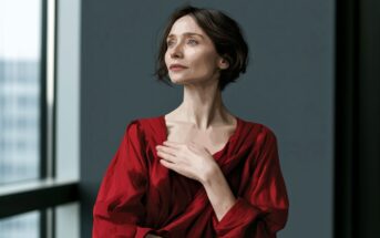 A woman in a red dress stands indoors by a large window, gazing thoughtfully to the side with one hand resting on her chest. The image has a painted, stylized effect.