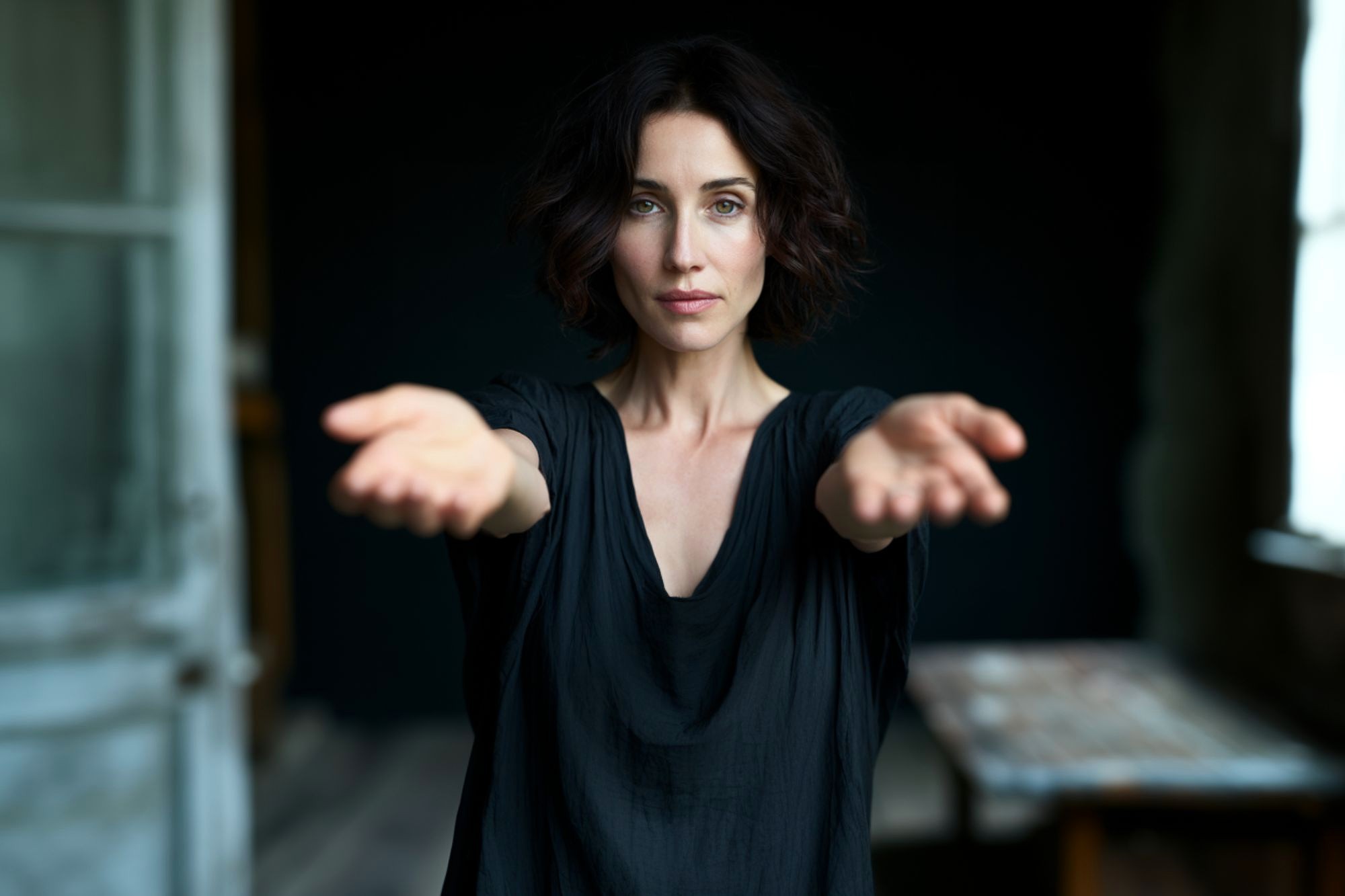 A woman with short dark hair stands indoors, facing forward with arms extended and palms up. She wears a loose black top and looks directly at the camera. The background is dimly lit and slightly blurred.