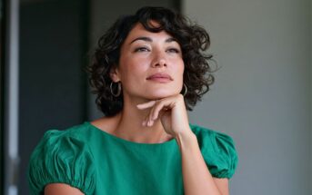 A woman with short, curly dark hair and a green top rests her chin on her hand while looking off to the side.