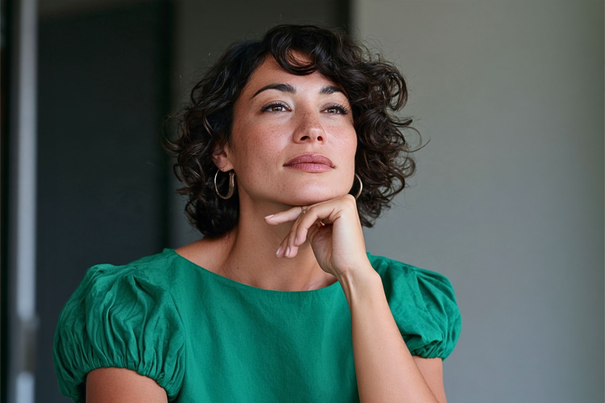 A woman with short, curly dark hair and a green top rests her chin on her hand while looking off to the side.