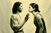 Two women face each other against a plain background; one appears calm, raising her hand as if to stop or gently reason, while the other leans forward, looking angry and confrontational.