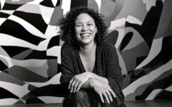 A smiling woman with curly hair kneeling in front of a black and white geometric patterned wall.