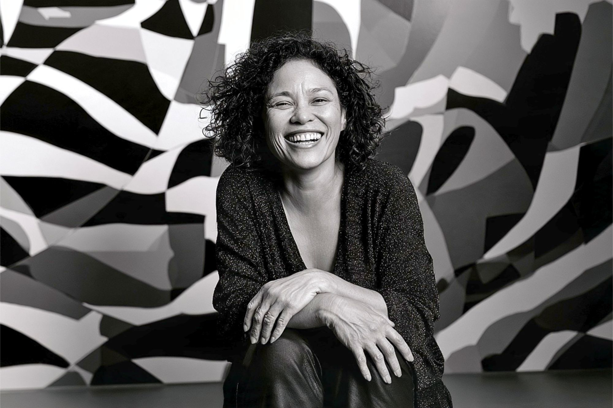 A smiling woman with curly hair kneeling in front of a black and white geometric patterned wall.
