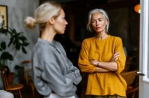 Two women stand indoors facing each other. The woman in the foreground, wearing gray, is out of focus, while the woman in the yellow sweater stands with arms crossed, looking serious or concerned. The background shows a cozy room.