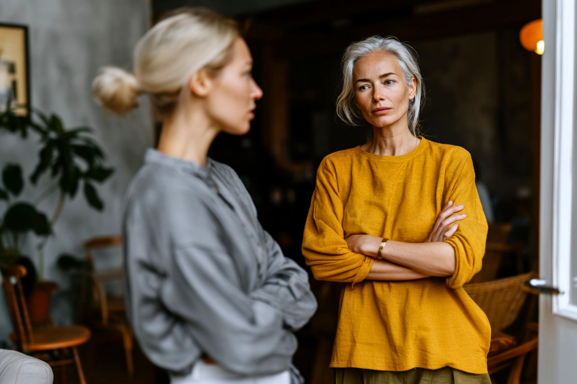 Two women stand indoors facing each other. The woman in the foreground, wearing gray, is out of focus, while the woman in the yellow sweater stands with arms crossed, looking serious or concerned. The background shows a cozy room.