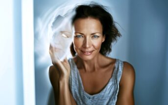 A woman with blue eyes and tousled hair holds a translucent, ghostly mask in front of her face, creating an ethereal effect. She wears a gray sleeveless top and stands in soft, cool lighting.
