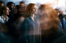 A woman with red hair stands still and looks serious in the middle of a blurred, moving crowd, creating a sense of isolation and contrast between her and her busy surroundings.