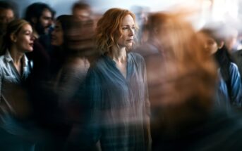 A woman with red hair stands still and looks serious in the middle of a blurred, moving crowd, creating a sense of isolation and contrast between her and her busy surroundings.
