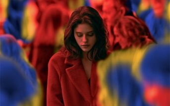 A woman in a red coat looks down, standing among a blurred crowd with vibrant red, blue, and yellow tones. The image conveys a feeling of isolation or introspection.