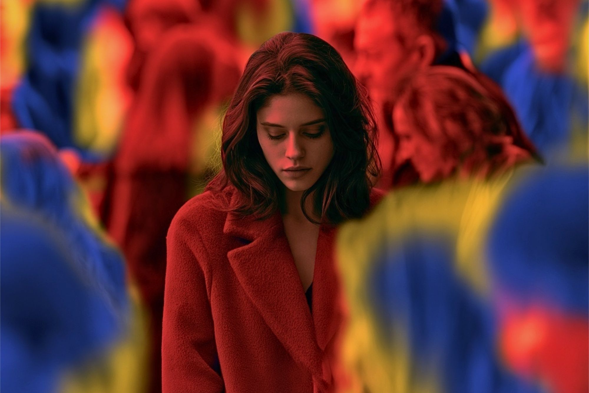 A woman in a red coat looks down, standing among a blurred crowd with vibrant red, blue, and yellow tones. The image conveys a feeling of isolation or introspection.