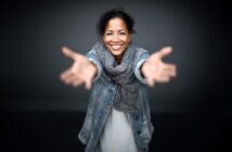 A woman smiling warmly, standing with arms outstretched toward the camera, wearing a denim jacket and a gray scarf against a dark background.