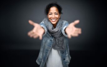 A woman smiling warmly, standing with arms outstretched toward the camera, wearing a denim jacket and a gray scarf against a dark background.