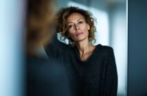 A woman with curly hair wearing a dark sweater leans against a reflective surface, looking directly at the camera with a thoughtful expression.