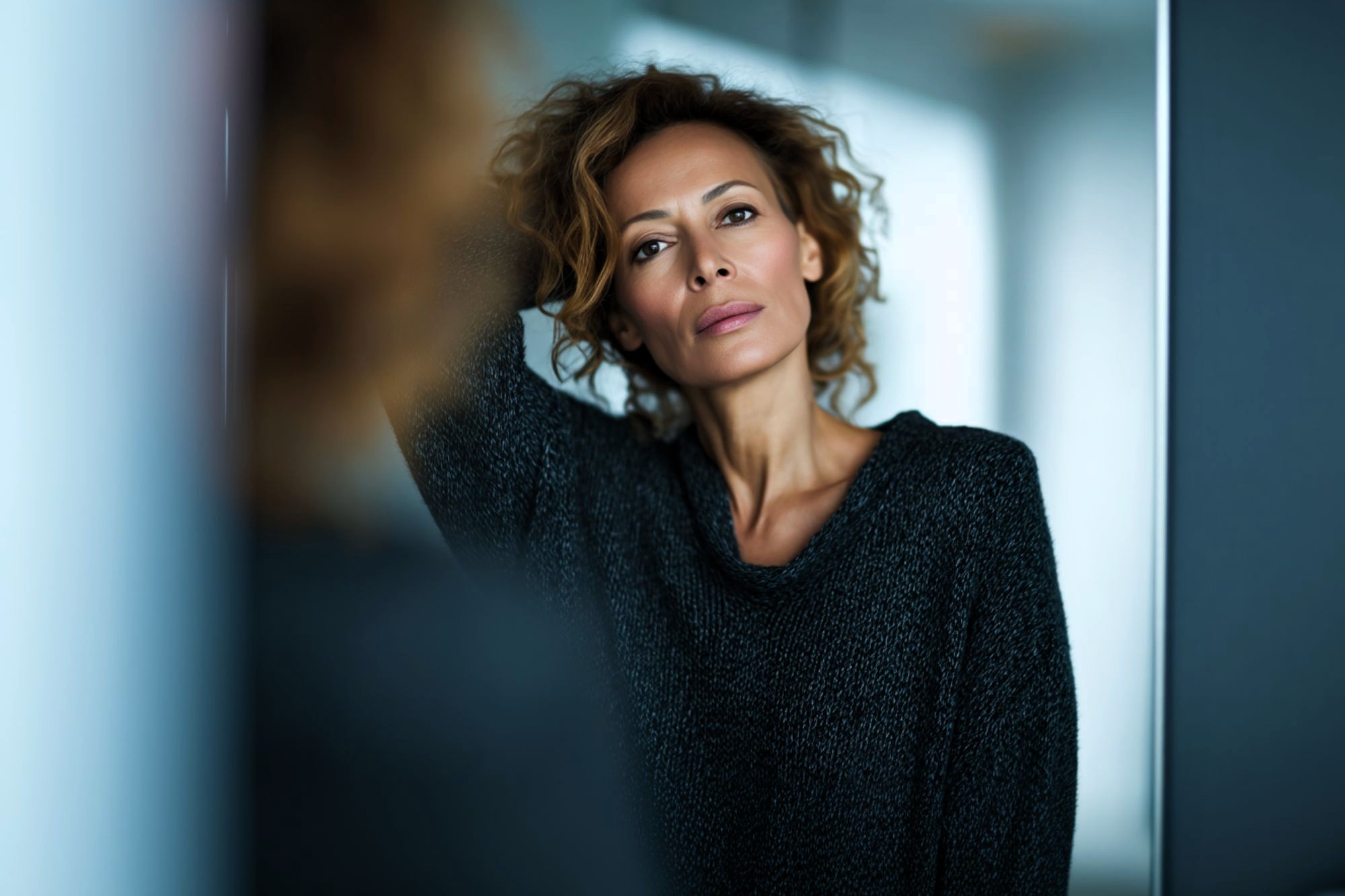 A woman with curly hair wearing a dark sweater leans against a reflective surface, looking directly at the camera with a thoughtful expression.