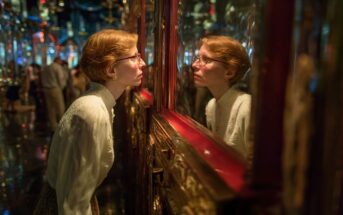 A woman with short red hair and glasses closely examines her reflection in an ornate mirror, surrounded by shimmering lights and intricate decorations.