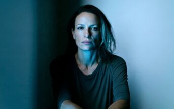 A woman with a serious expression looking directly at the camera in blue-toned lighting.