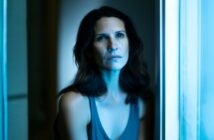 A woman with wavy brown hair sits by a window, illuminated by cool blue light, looking slightly upward with a serious, contemplative expression. She wears a sleeveless top and is framed by soft-focus surroundings.