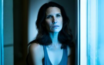 A woman with wavy brown hair sits by a window, illuminated by cool blue light, looking slightly upward with a serious, contemplative expression. She wears a sleeveless top and is framed by soft-focus surroundings.